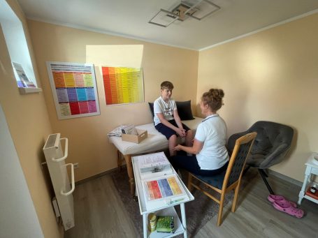 Eine Frau behandelt ein Kind in einem hellen, freundlichen Raum. Farbige Grafiken hängen an der Wand.