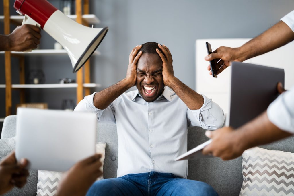 Mann sitzt verzweifelt auf dem Sofa, umgeben von Menschen, die ihm Dokumente und einen Lautsprecher zeigen.