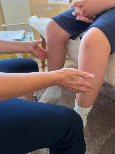 Eine Person untersucht das Bein eines anderen, beide sitzen auf einem Sofa.