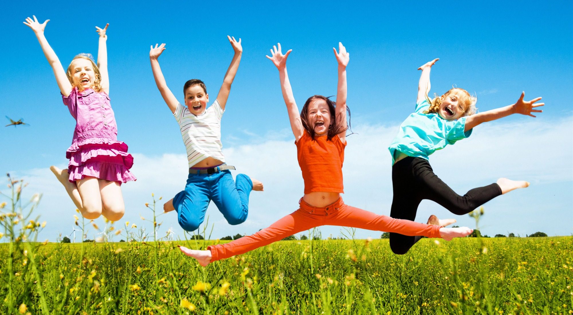Fünf fröhliche Kinder springen in einer blühenden Wiese unter blauem Himmel.