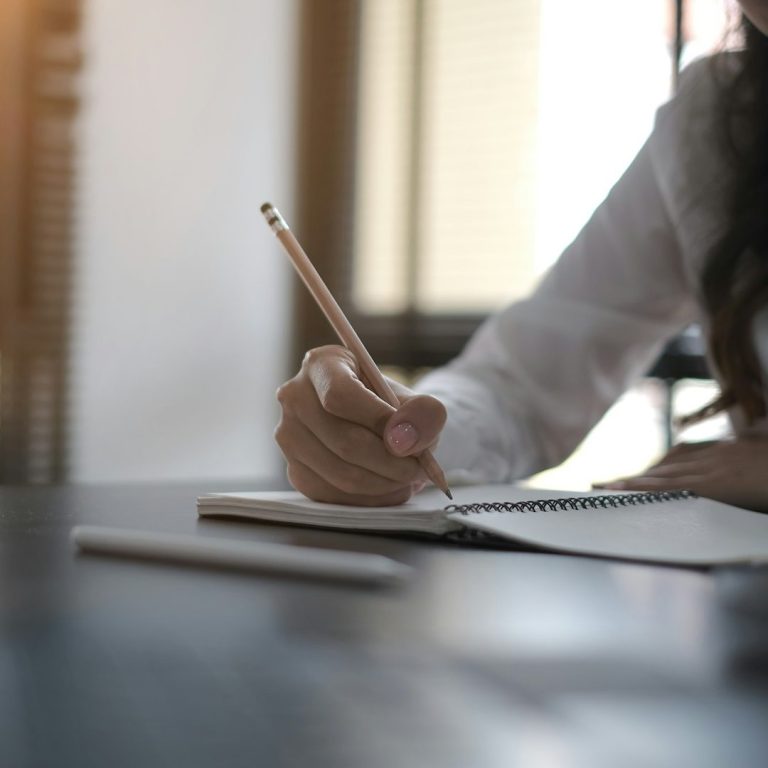 Eine Hand hält einen Bleistift und schreibt in ein Notizbuch auf einem Tisch.