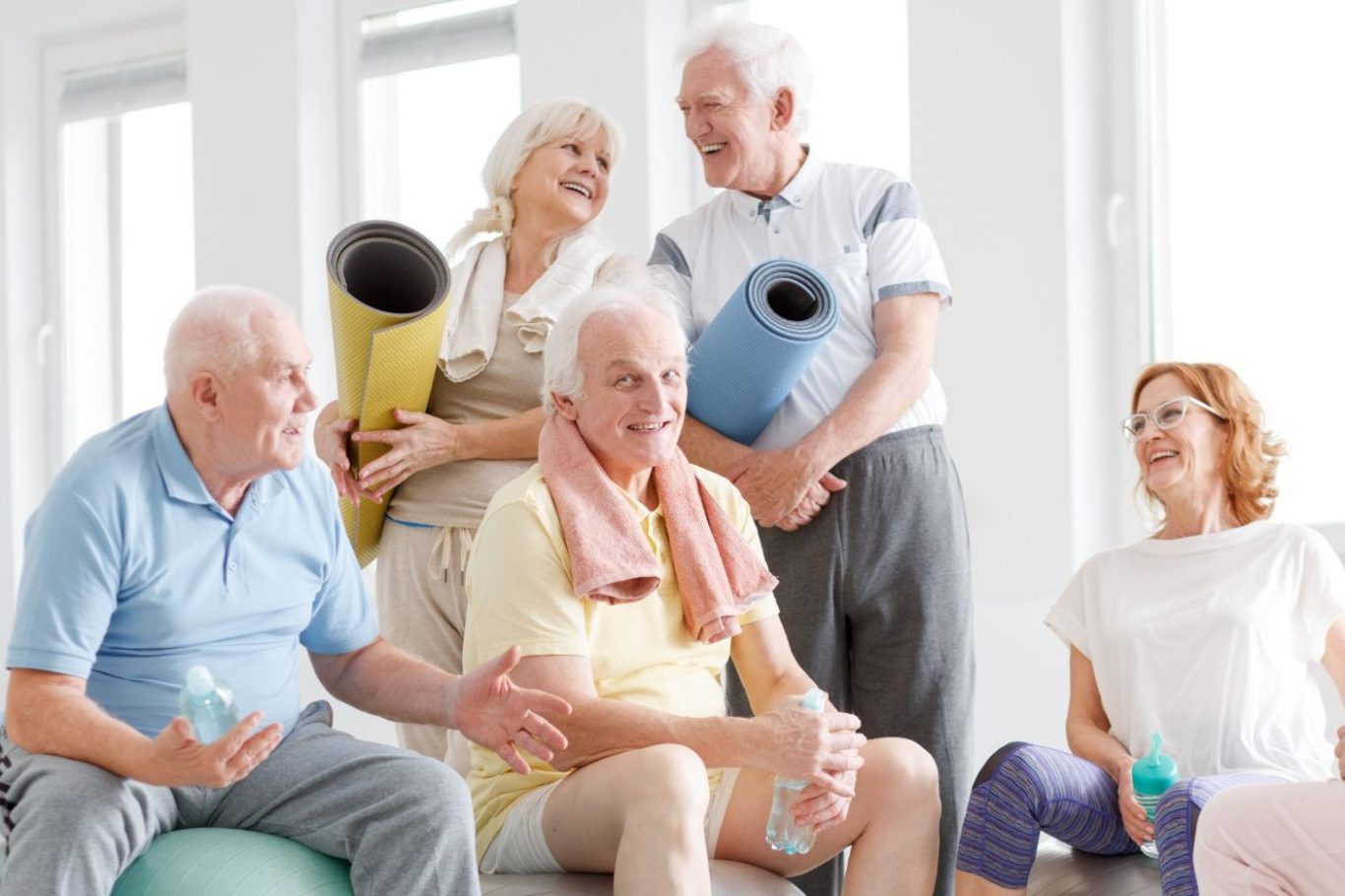 Ältere Menschen in einer Fitnessgruppe, lachen und halten Yogamatten und Wasserflaschen.
